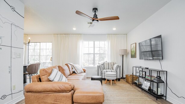 A living room with a brown leather sofa, a striped armchair, a round ottoman, a floor lamp, a TV mounted on the wall, and a large window with curtains. - Downtown Trailside Cottage (Bentonville)