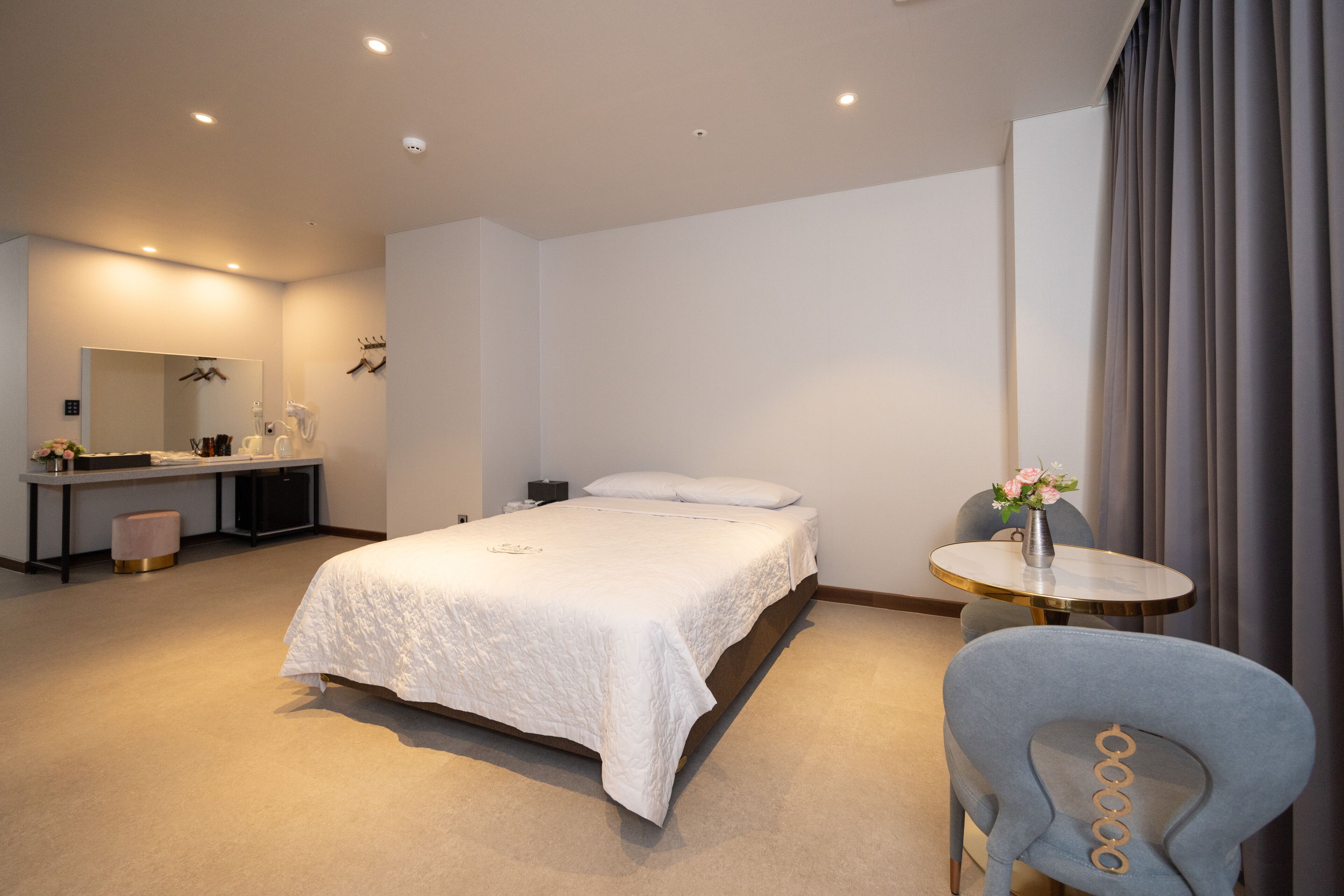 A modern hotel room with a large bed, a desk with a mirror, a chair, and a small table with a vase of flowers.