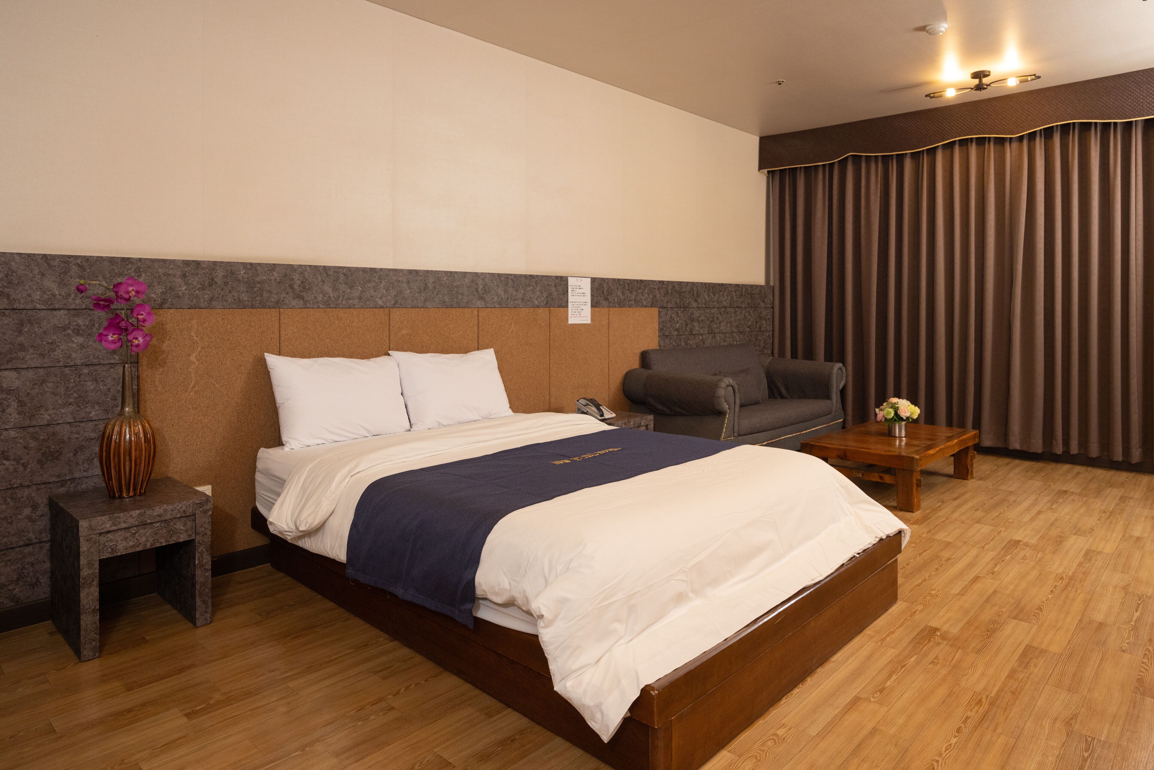Habitación de hotel con una cama de madera, una mesita de noche con un florero, un sillón gris y una mesa de centro de madera.