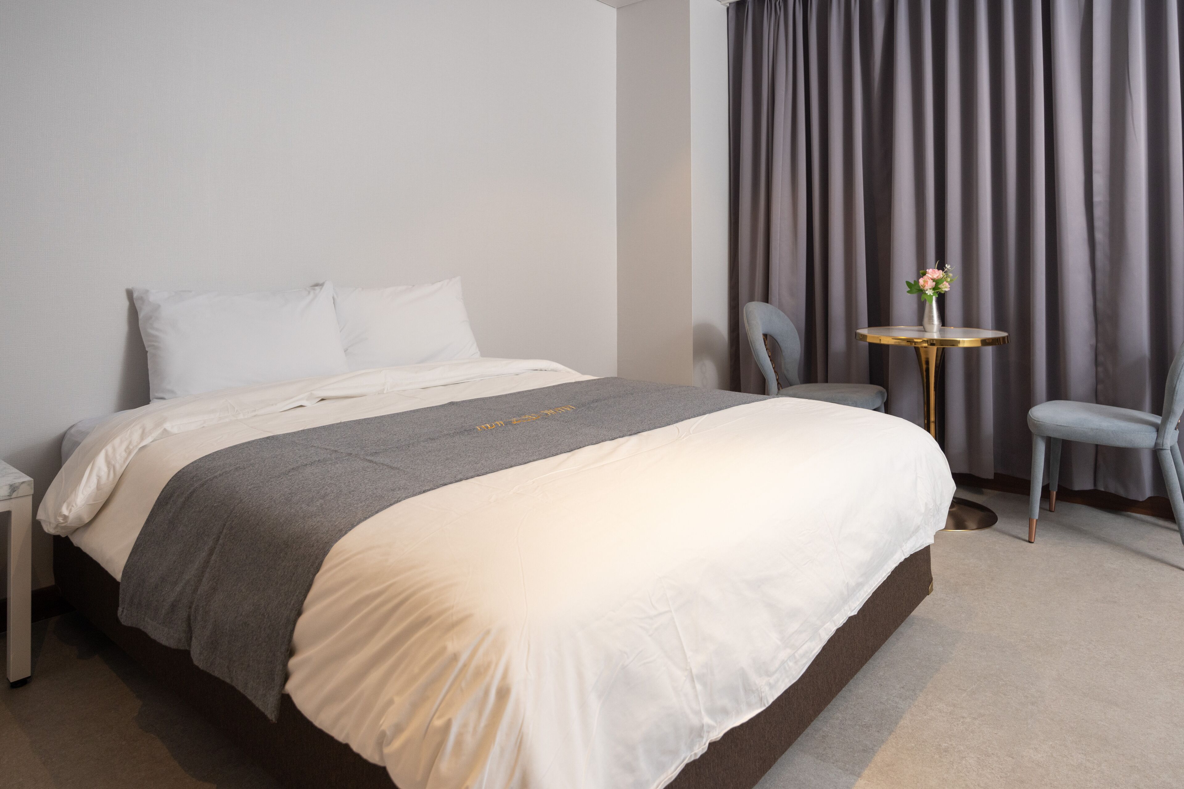 Una cama bien hecha con edredón gris y blanco, una mesita redonda con un florero y una silla gris en una habitación de hotel moderna.