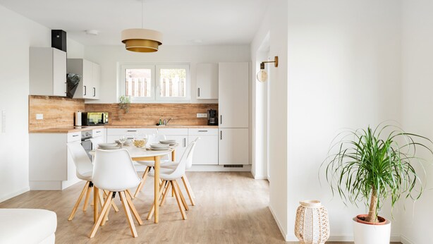 A modern kitchen with white cabinets, a wooden countertop, and a dining area with white chairs. - Ferienwohnung Zur Örtze (Faßberg)