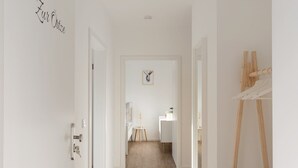 A hallway with a wooden floor, white walls, and a door on the left. There is a small table with a plant and a lamp in the background. - Ferienwohnung Zur Örtze (Faßberg)