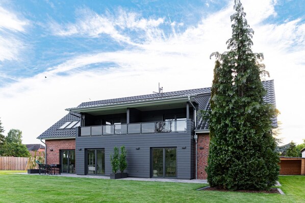 Une maison moderne à deux étages, dotée d’un balcon, d’une pelouse bien entretenue et d’un grand arbre à feuilles persistantes.