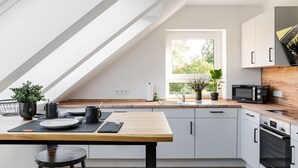 A modern kitchen with a wooden table, white cabinets, a built-in oven, and a window with a view of greenery. - Ferienwohnung Heideloft (Faßberg)
