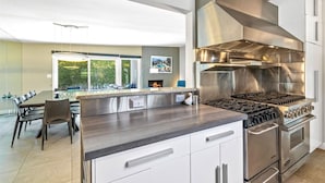 Una cocina moderna con electrodomésticos de acero inoxidable, una zona de comedor con una mesa grande y sillas, y vistas a zonas verdes por las ventanas.