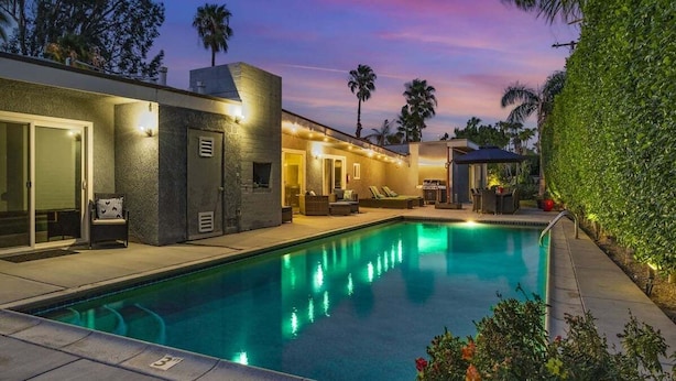 Una piscina con zona de patio, espacio de descanso cubierto y vistas a palmeras.