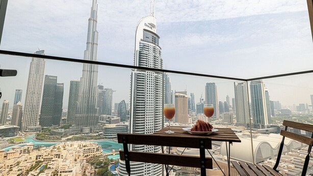 A balcony with a table, two glasses of a beverage, and a croissant. The view includes a cityscape with several high-rise buildings, including the Burj Khalifa.