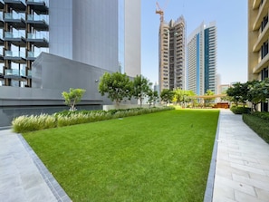 A well-maintained grassy area with a paved walkway, surrounded by modern buildings and construction cranes in the background.