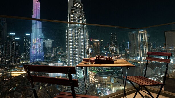 A balcony with a table and two chairs, overlooking a cityscape at night with illuminated buildings, including a prominent tower with 'EMAR' signage.