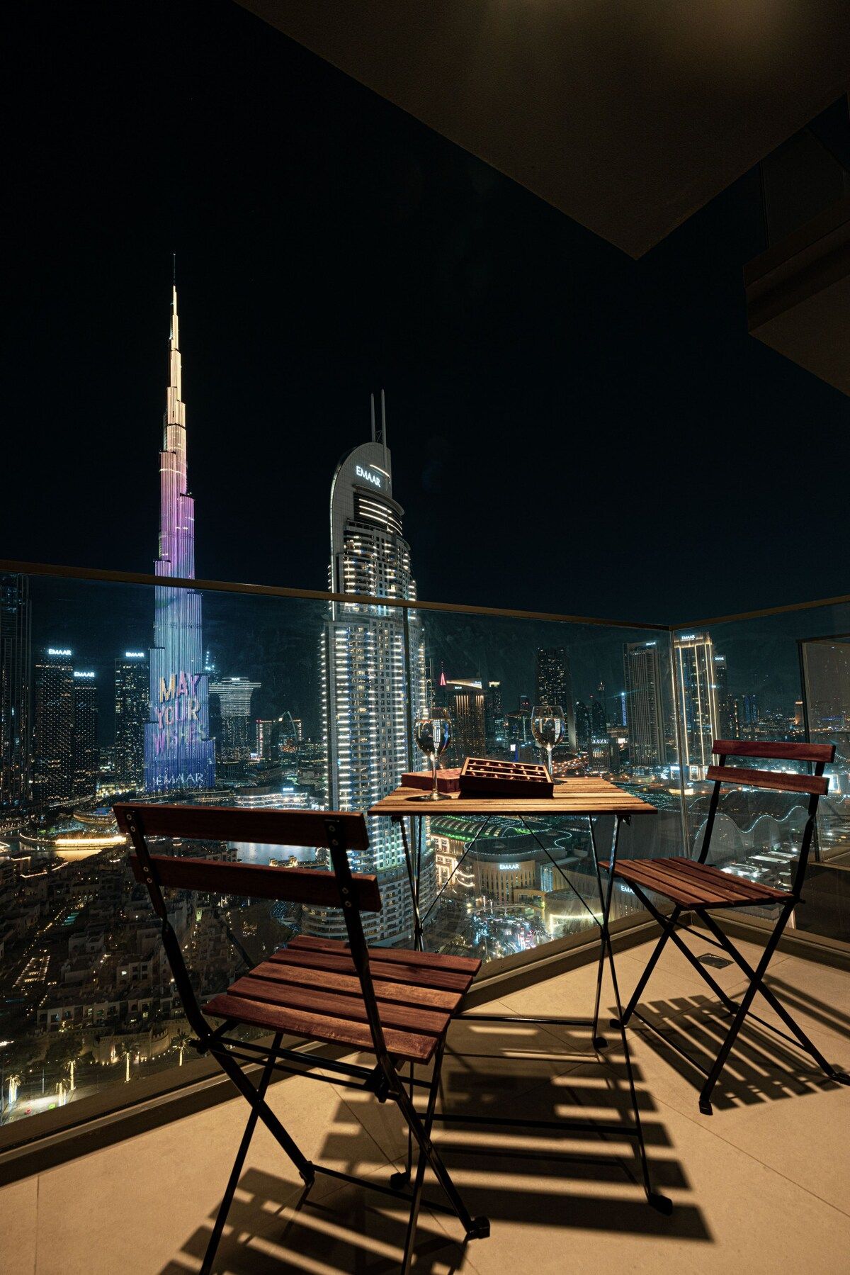 Un balcon avec deux chaises en bois et une petite table, d’où l’on peut admirer la vue sur la ville la nuit, avec ses bâtiments illuminés.