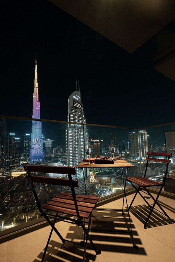 A balcony with two wooden chairs and a small table, overlooking a cityscape at night with illuminated buildings.