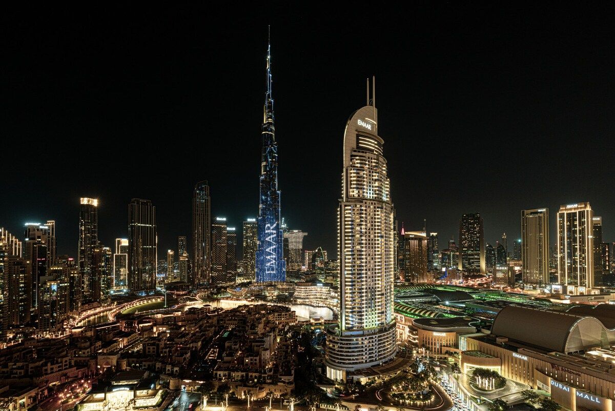 A nighttime cityscape with prominent skyscrapers, including a tall, illuminated tower and a curved building with 'EMAR' signage.