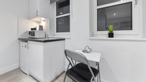 A compact kitchen with white cabinetry, a microwave, and a small island with a sink. There is a window with a plant on the windowsill and a chair at a small table. - Serviced Ensuite Room Crystal Palace London Se20 (London)