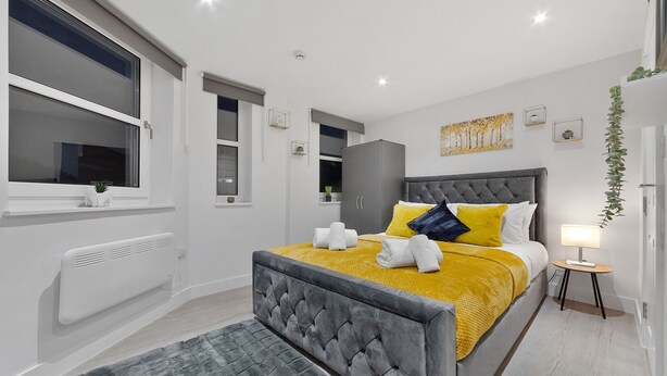 A modern bedroom with a large bed, a grey upholstered headboard, and a yellow throw blanket. - Serviced Ensuite Room Crystal Palace London Se20 (London)