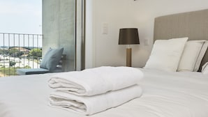 A neatly made bed with white linens and a folded towel, a bedside lamp, and a view of the outdoors through a large window. - Porto Ocean Flat (Vila Nova de Gaia)