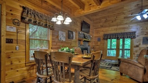 A wooden cabin interior with a dining table, four chairs, a fireplace, and a large window. - Welcome to Relaxing River Retreat, where your pets are welcome amidst a scenic riverfront, screened porch, hot tub, WiFi, firepit, and abundant wildlife (Ellijay)