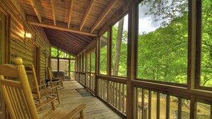 A covered wooden deck with benches, large windows, and a view of a forest. - Welcome to Relaxing River Retreat, where your pets are welcome amidst a scenic riverfront, screened porch, hot tub, WiFi, firepit, and abundant wildlife (Ellijay)