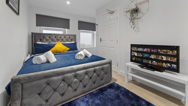 A modern bedroom with a grey tufted headboard, a blue bedspread, and a flat-screen TV on a white stand.