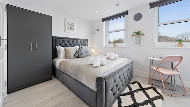Quarto moderno com uma cama grande, um guarda-roupa cinzento, uma escrivaninha com cadeira e uma janela com vista para o exterior.