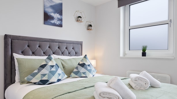 Ein ordentlich gemachtes Bett mit grauem Kopfteil, grüner Bettwäsche und zwei geometrisch gemusterten Kissen. Auf dem Bett liegen gerollte weiße Handtücher, und ein Fenster bietet Blick auf den Himmel, während ein kleiner Topf mit einer Pflanze auf der Fensterbank steht.