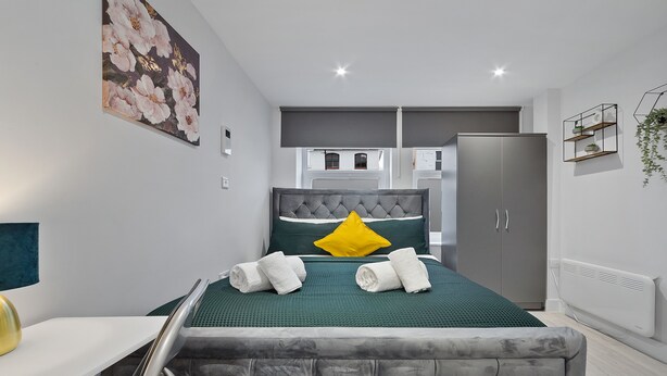 Ein modernes Schlafzimmer mit grauem, gepolstertem Kopfteil, einer grünen Tagesdecke und einem gelben Kissen. An der Wand befindet sich ein weißer Heizkörper und ein grauer Kleiderschrank.