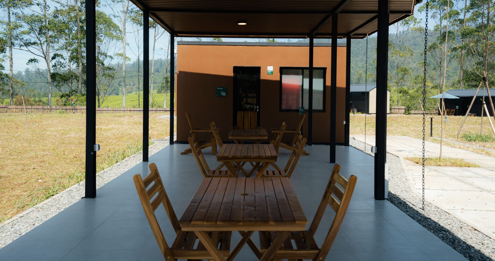 An outdoor covered dining area with wooden tables and chairs, a view of a natural landscape, and a building in the background.