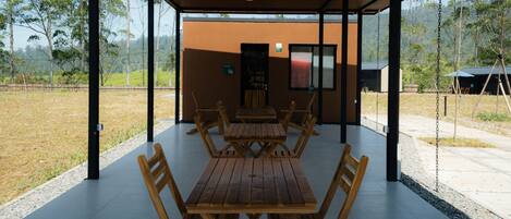 An outdoor covered dining area with wooden tables and chairs, a view of a natural landscape, and a building in the background.