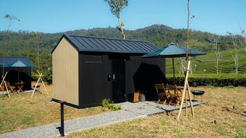 Una cabaña pequeña de color oscuro con techo inclinado, una puerta y una ventana, rodeada por un sendero de grava y sillas de madera.