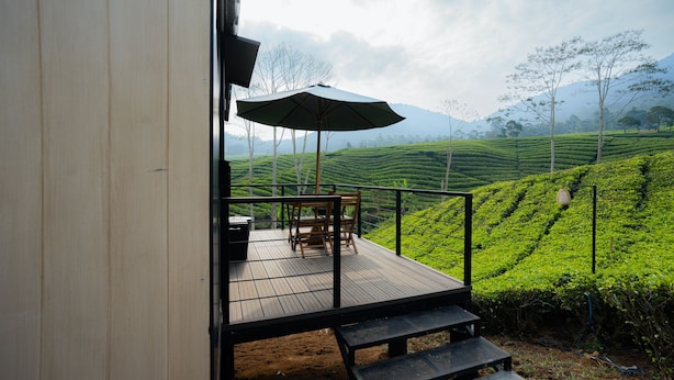 A wooden deck with a table and chairs, an umbrella, and a view of tea plantations.