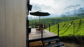 A wooden deck with a table and chairs, an umbrella, and a view of tea plantations.