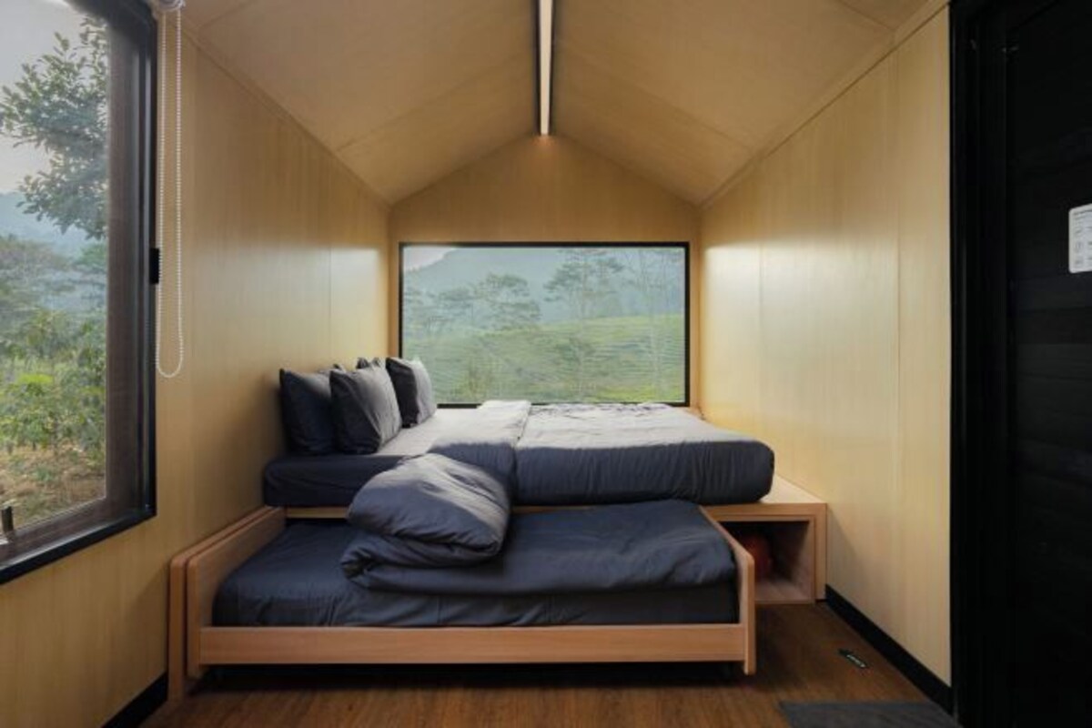 A compact, modern bedroom with a bed, pillows, and a large window offering a view of greenery.