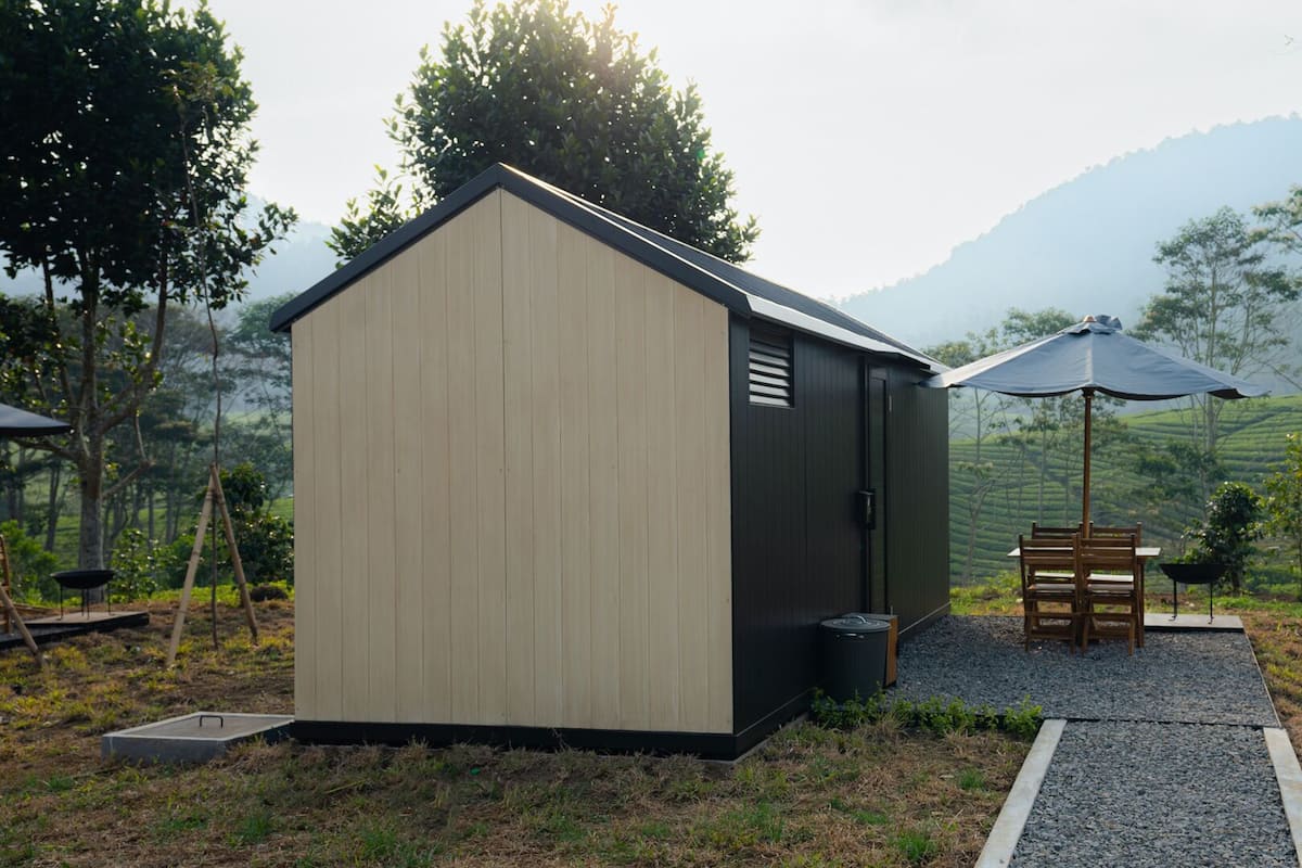 Een klein, laag gebouw met een schuin dak, een deur en een raam. Buiten staan een tafel en stoelen, en een zwarte prullenbak is zichtbaar. Het gebouw ligt aan een grindpad, omringd door groen, bomen en een heuvelachtig landschap op de achtergrond.