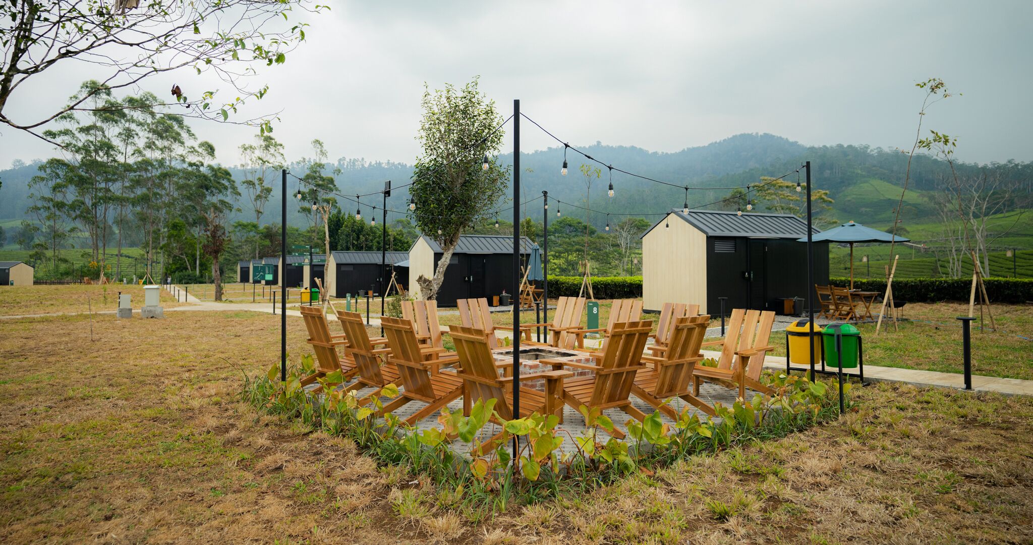 A circular seating area with wooden chairs around a central fire pit, surrounded by a grassy field with a backdrop of hills and trees.
