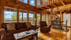 Eine geräumige Holzhütte mit großem Sofa, Sesseln und Esstisch, ausgestattet mit großen Fenstern und einem Balkon.