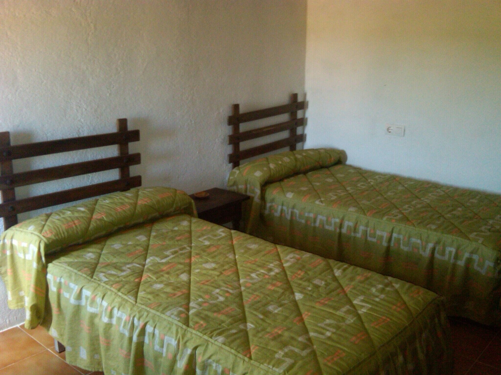 Una habitación con dos camas individuales con cabeceros de madera, una mesita de noche pequeña y una pared sin decorar.