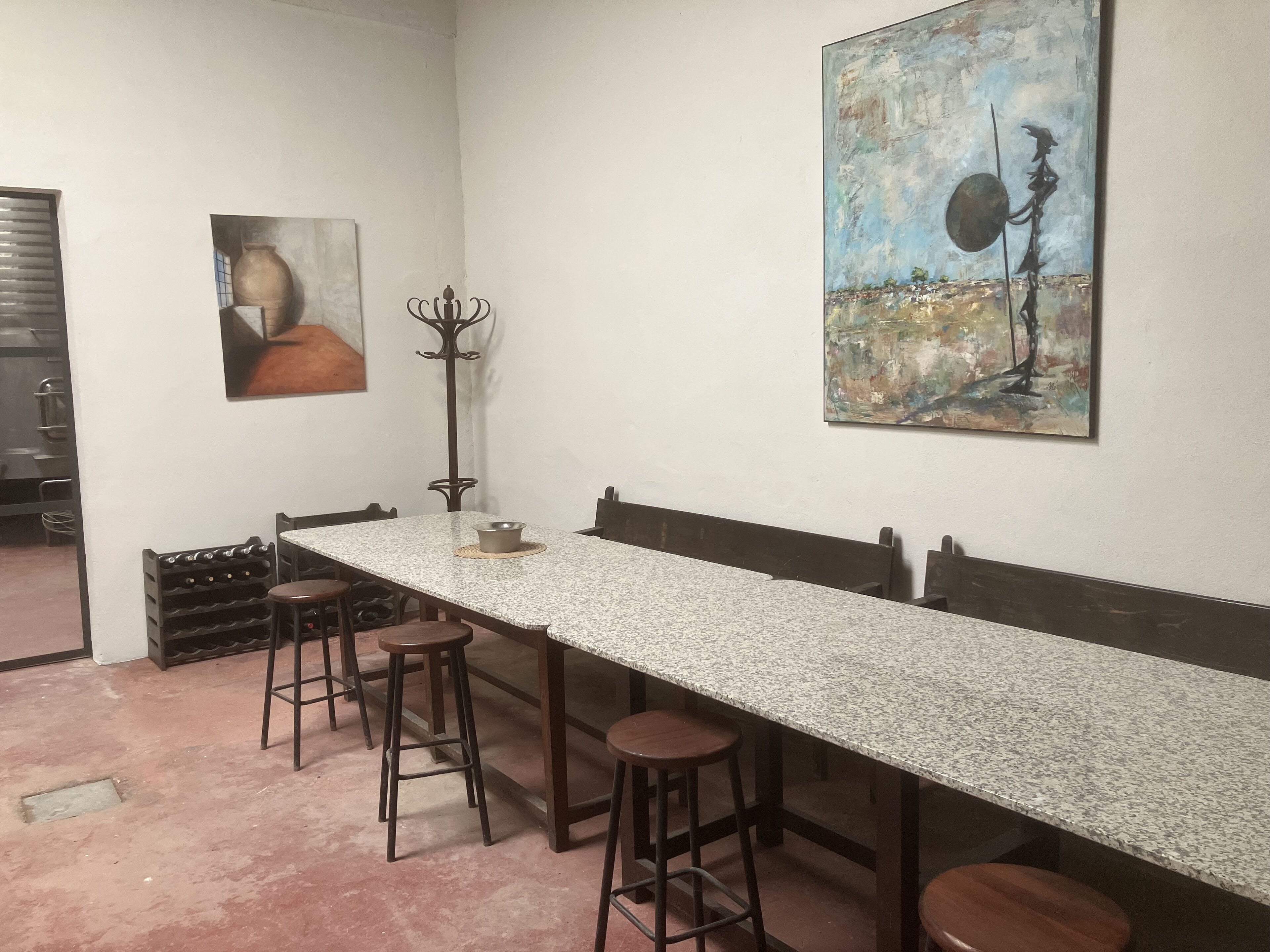 Une longue table à manger, quatre tabourets, un présentoir en métal et deux tableaux accrochés au mur.
