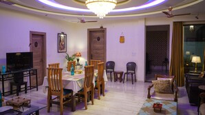 A dining area with a chandelier, wooden chairs, a table with a tablecloth, and a television on a stand. - Wild Pensione Resort  (Ramnagar)