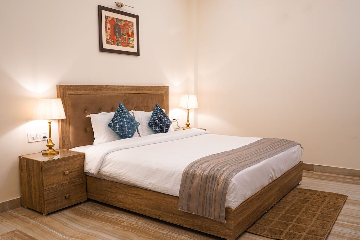 A neatly made bed with a wooden headboard, two bedside lamps, a small wooden nightstand with a drawer, and a patterned throw blanket.