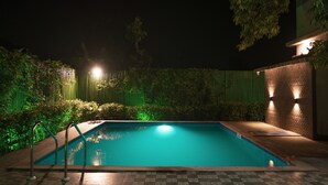A swimming pool at night with surrounding greenery and a brick wall with lights. - Wild Pensione Resort  (Ramnagar)