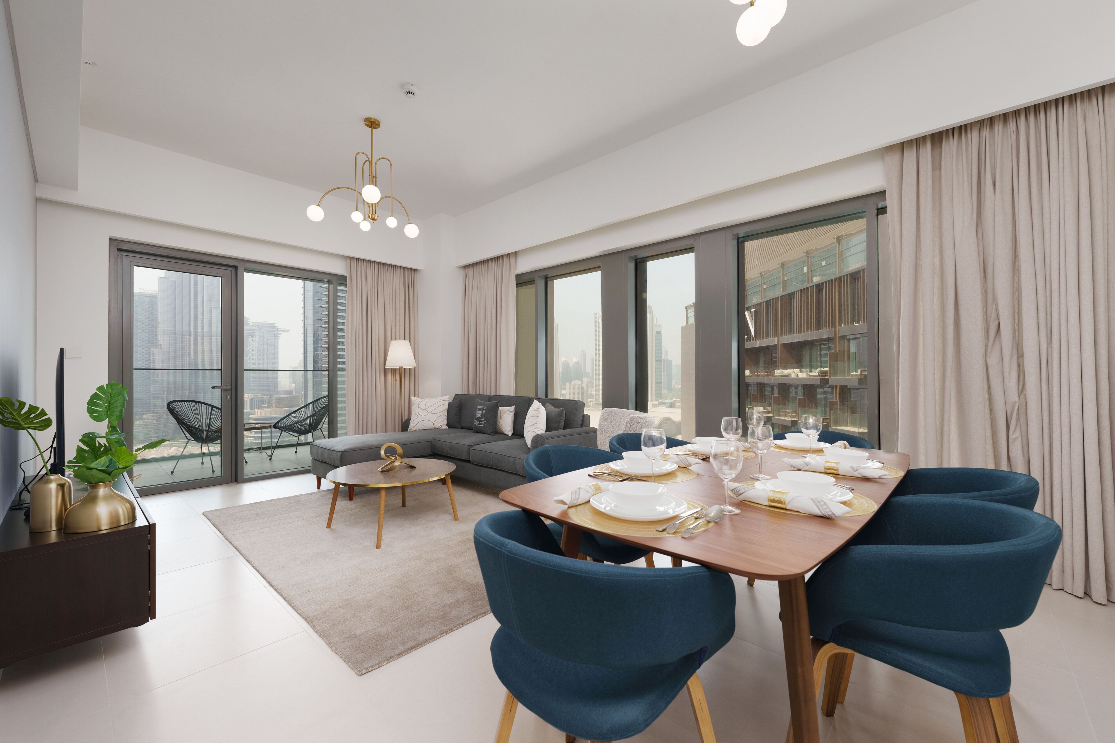 A modern dining area with a wooden table set for six, surrounded by blue chairs, a grey sofa, and a small round coffee table. Large windows offer a view of the cityscape.