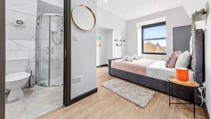 Un dormitorio moderno con ducha de vidrio, cama con cabecera gris y un ventanal grande.
