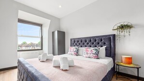 Un dormitorio con una cama grande, una ventana, un radiador, una mesita de noche con una lámpara y una planta colgante.