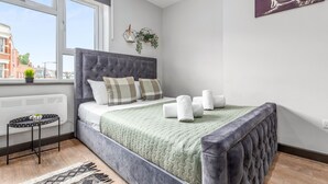 A bedroom with a large bed, a window, a radiator, a small table, and a wall-mounted artwork. - Cozy Serviced En-suite Room Sydenham London Se26 (London)