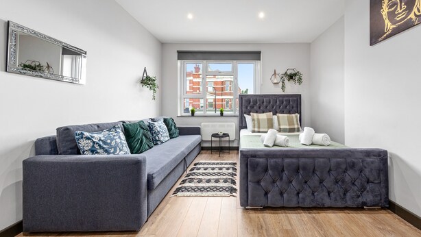A modern living room with a grey sofa, a tufted ottoman, a small table, and a window with a view of buildings. - Cozy Serviced En-suite Room Sydenham London Se26 (London)