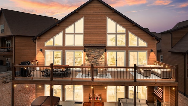 Una casa de dos pisos con un balcón grande, chimenea de piedra y varias ventanas.