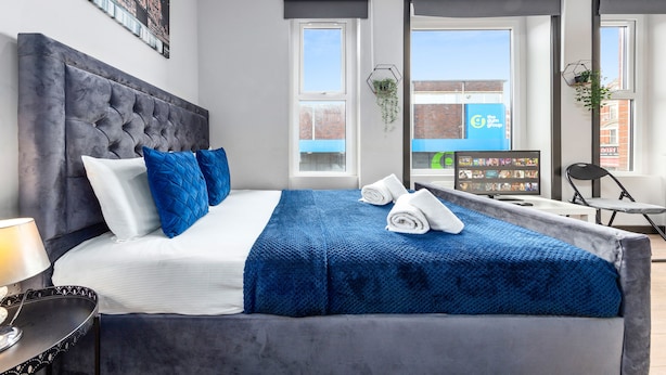 Una habitación de hotel moderna con una cama grande, un cabecero tapizado en gris y vistas de la ciudad a través de amplias ventanas.