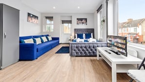 A modern bedroom with a grey tufted headboard, a blue sofa, and a TV on a white desk. - Sleep 4 Serviced Ensuite Room Sydenham London Se26 (London)