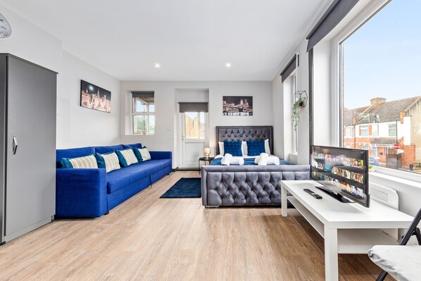 A modern bedroom with a grey tufted headboard, a blue sofa, and a TV on a white desk.