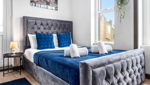 Un dormitorio con cabecera capitonada, cama con ropa de cama azul y blanca, una manta azul y una planta colgante.