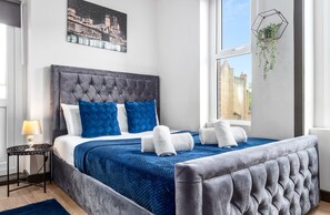 A bedroom with a tufted headboard, a bed with blue and white bedding, a blue throw blanket, and a hanging plant. - Sleep 4 Serviced Ensuite Room Sydenham London Se26 (London)